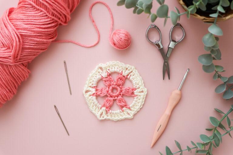 Crochet flower hexagon motif in pink and cream yarn, displayed on a pink background with crochet tools including a peach ergonomic hook, scissors, yarn needles, and eucalyptus leaves.