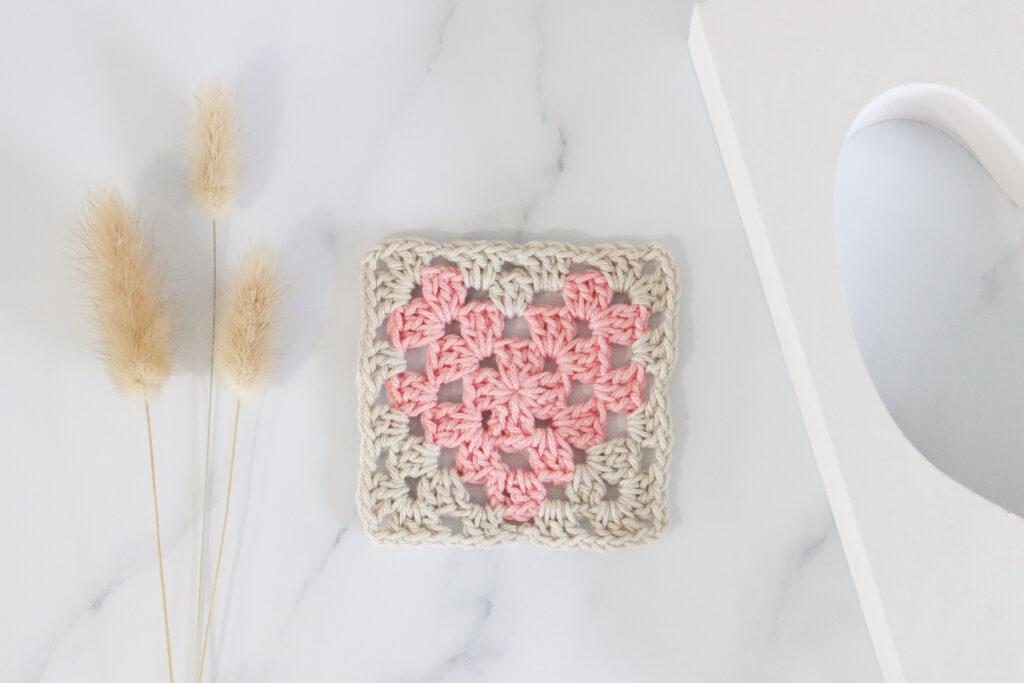 a flay lay of a heart granny square made in two colours, the heart is formed using a pale pink yarn and beige yarn used for the border. The granny sqaure is lay on a marble backdrop with dried flowers lay to the left of the swatch.