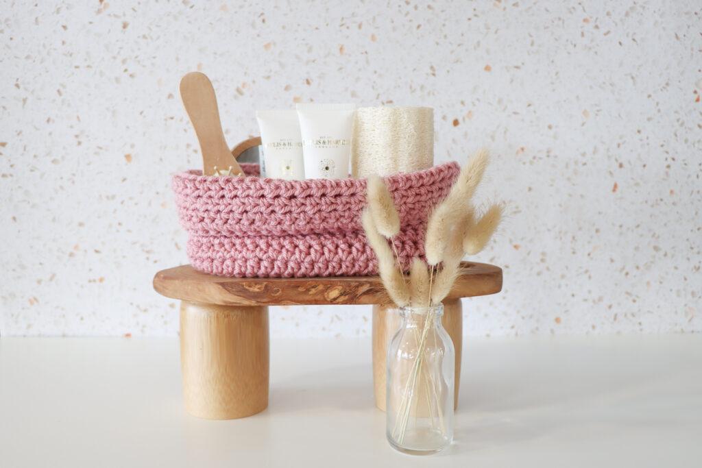 Oval crochet basket with self care goodies inside, surrounded by a vase of botanicals on a wooden table.