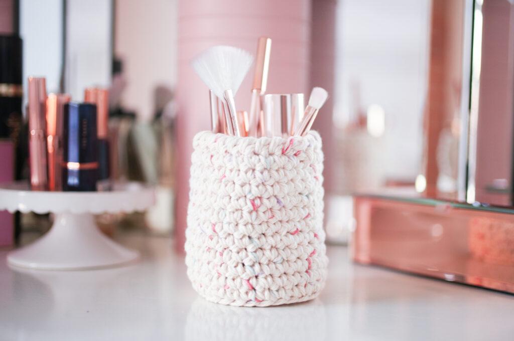 A crochet nesting basket in a beauty setting filled with make up brushes on a dressing table.