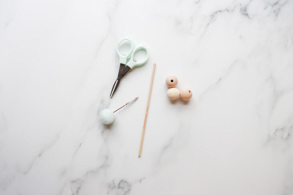 A group of crochet tools including silver and mint embroidery scissors, a wooden crochet hook, 3 wooden beads and a darning needle inserted into a mink coloured felt ball.