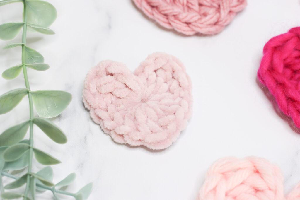 Crochet heart made in light pink chenille yarn laid on a marble background with a hint of green foliage and other crochet hearts
