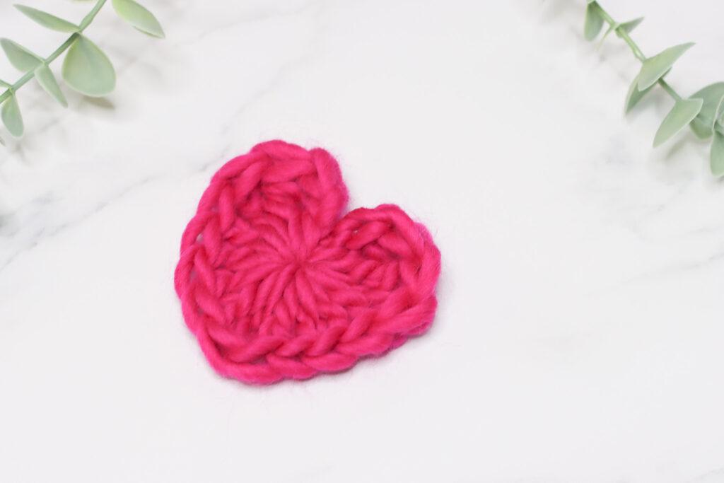 Crochet heart made in light pink chunky yarn laid on a marble background with a hint of green foliage