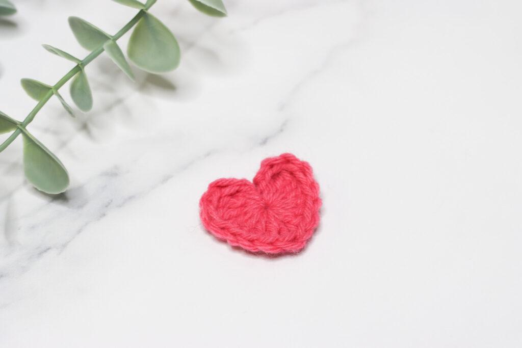 Crochet heart made in dark pink yarn laid on a marble background with a hint of green foliage