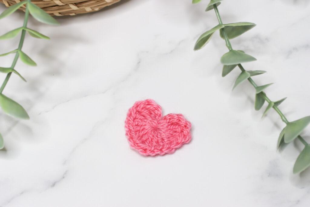Crochet heart made in pink yarn laid on a marble background with a hint of green foliage
