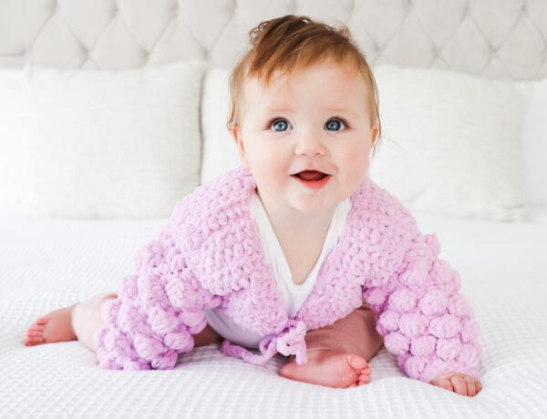 baby girl sat on a white bed wearing a textured baby bolero in lilac yarn.
