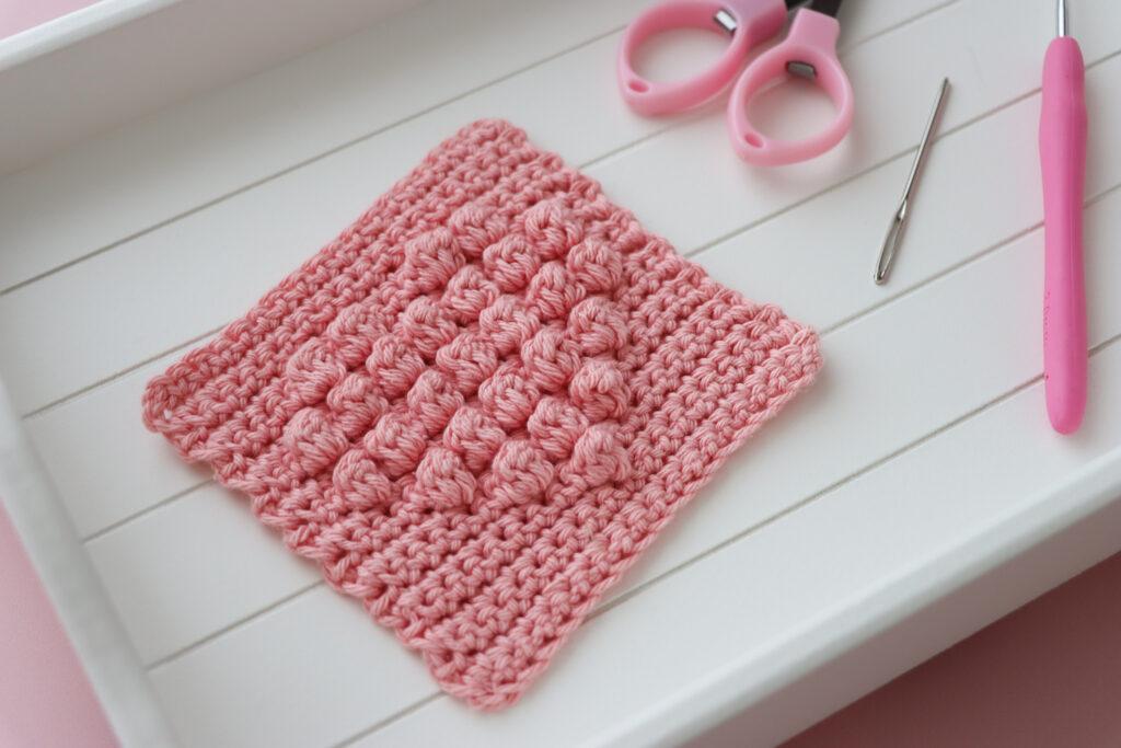 a crochet bobble heart square on a white wooden tray alongside crafting scissors and a darning needle.