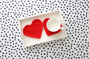 Two red crochet heart coasters on a white wooden tray. One with a mug on.