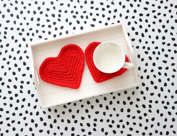 Two red crochet heart coasters on a white wooden tray. One with a mug on.