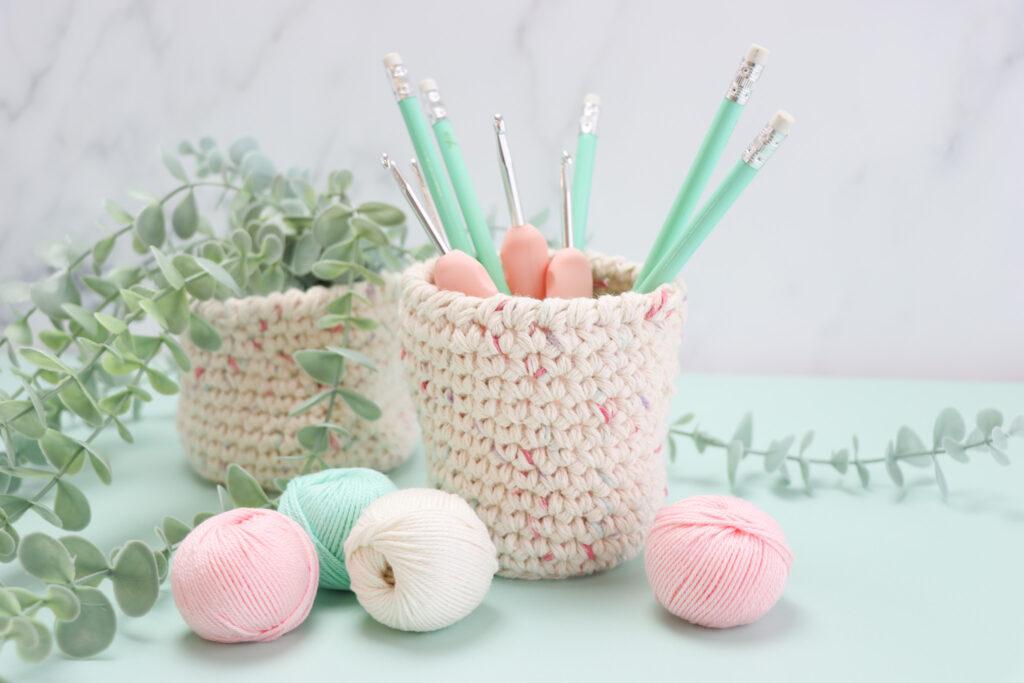 Two crochet nesting baskets with pencils and crochet hooks in surrounded by a plant and balls of yarn.