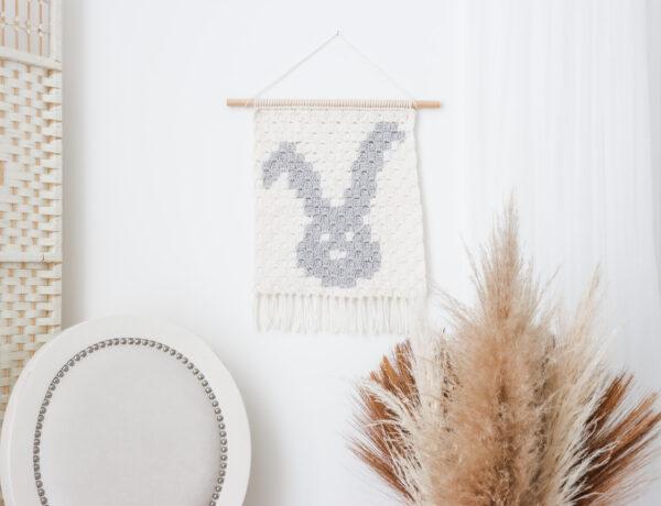 A bunny wall hanging using the corner to corner crochet method on a white wall with plants and a chair surrounding it.