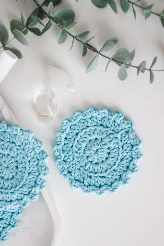 flat lay image of  blue face scrubbies on a white background with green foliage at the top of the image.