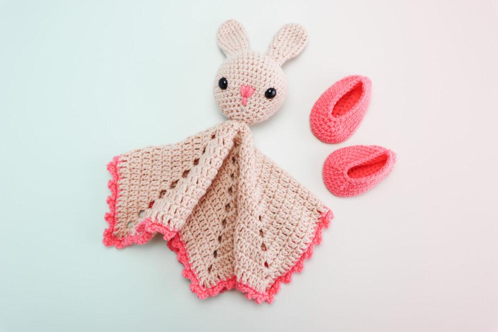 a crochet bunny lovely made in cream and pink yarn lay flat with a pair of pink crochet baby booties lay to the right of the bunny lovely on a ombre backdrop pale blue going into pink