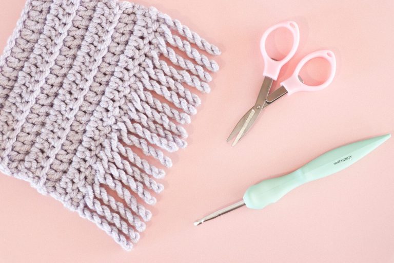 Close-up of a crochet project with twisted fringe in soft lilac yarn, pink-handled embroidery scissors, and a mint green ergonomic crochet hook on a pastel pink background.