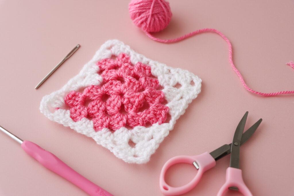 a heart granny square with yarn, scissors and a darning needle lay flat alongside on a pink background 