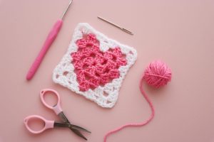 a heart granny square with yarn, scissors and a darning needle lay flat alongside on a pink background