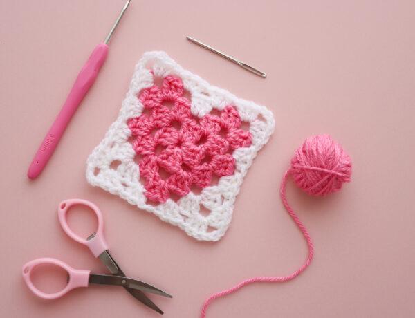 a heart granny square with yarn, scissors and a darning needle lay flat alongside on a pink background