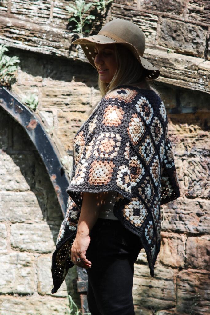 female with should length blonde hair stood in front of a stone bridge wearing a crochet poncho made of granny squares in a variety of colours including: dark grey, cream, camel and brown wearing a camel coloured hat and black skinny jeans. 