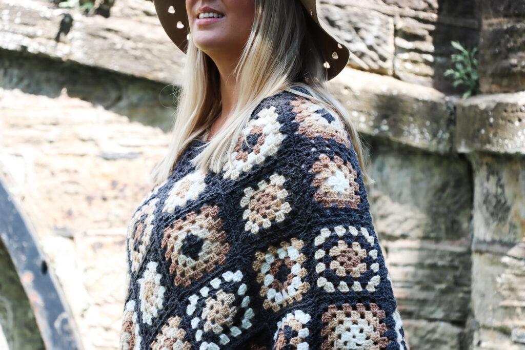 female with should length blonde hair stood in front of a stone bridge wearing a crochet poncho made of granny squares in a variety of colours including: dark grey, cream, camel and brown wearing a camel coloured hat and black skinny jeans.