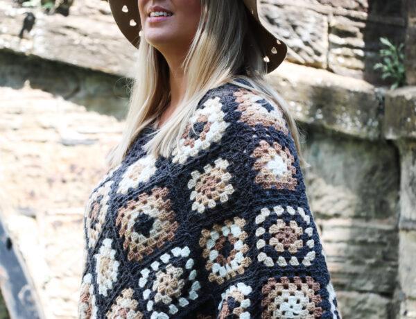 female with should length blonde hair stood in front of a stone bridge wearing a crochet poncho made of granny squares in a variety of colours including: dark grey, cream, camel and brown wearing a camel coloured hat and black skinny jeans.