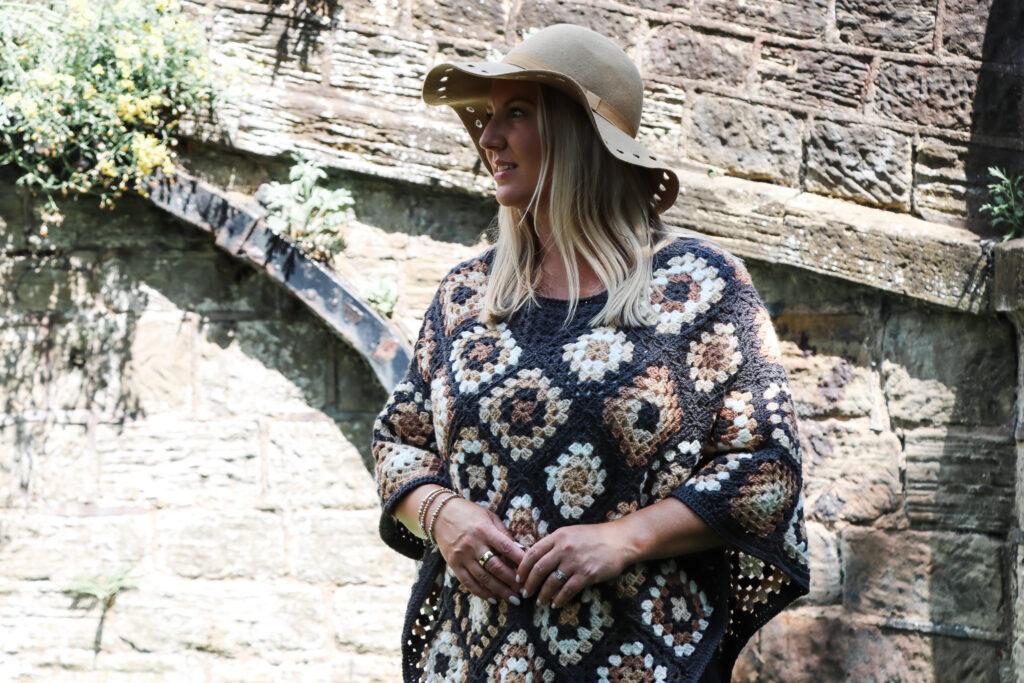 female with should length blonde hair stood in front of a stone bridge wearing a crochet poncho made of granny squares in a variety of colours including: dark grey, cream, camel and brown wearing a camel coloured hat and black skinny jeans. 