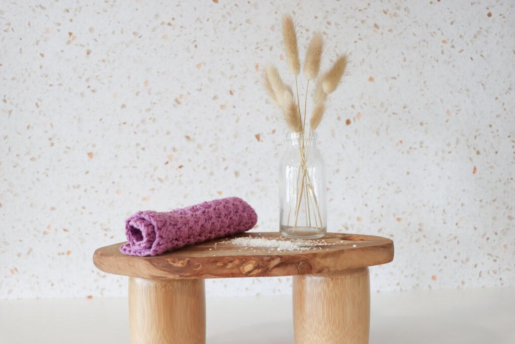 A purple crocheted wash cloth on a wooden table with a vase of botanicals.
