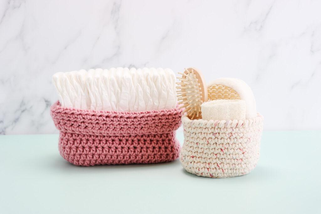 two crochet nesting basket sat next to each other one oval nesting basket filled with nappies and the second a small taller nesting basket filled with self care items