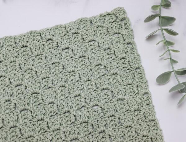crochet swatch of the corner to corner stitch in a pale green yarn lay flat on a white marble backdrop with foliage in the top right corner of the image.