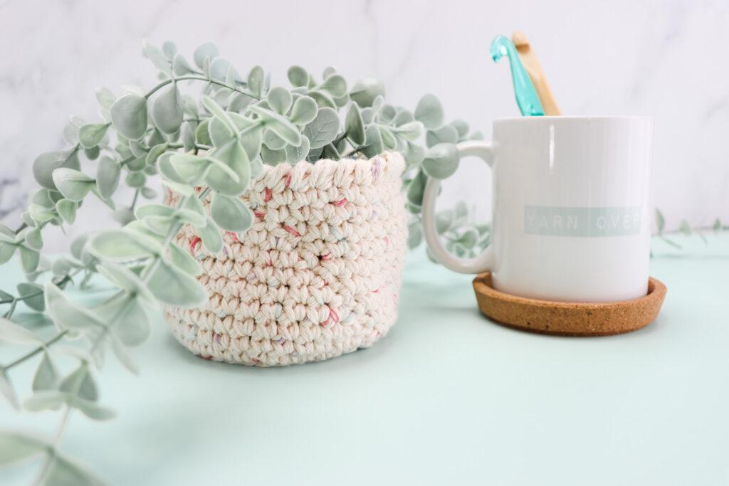 A crochet nesting basket with a plant in next to a mug with 'Yarn over' on it and 2 hooks inside the mug.