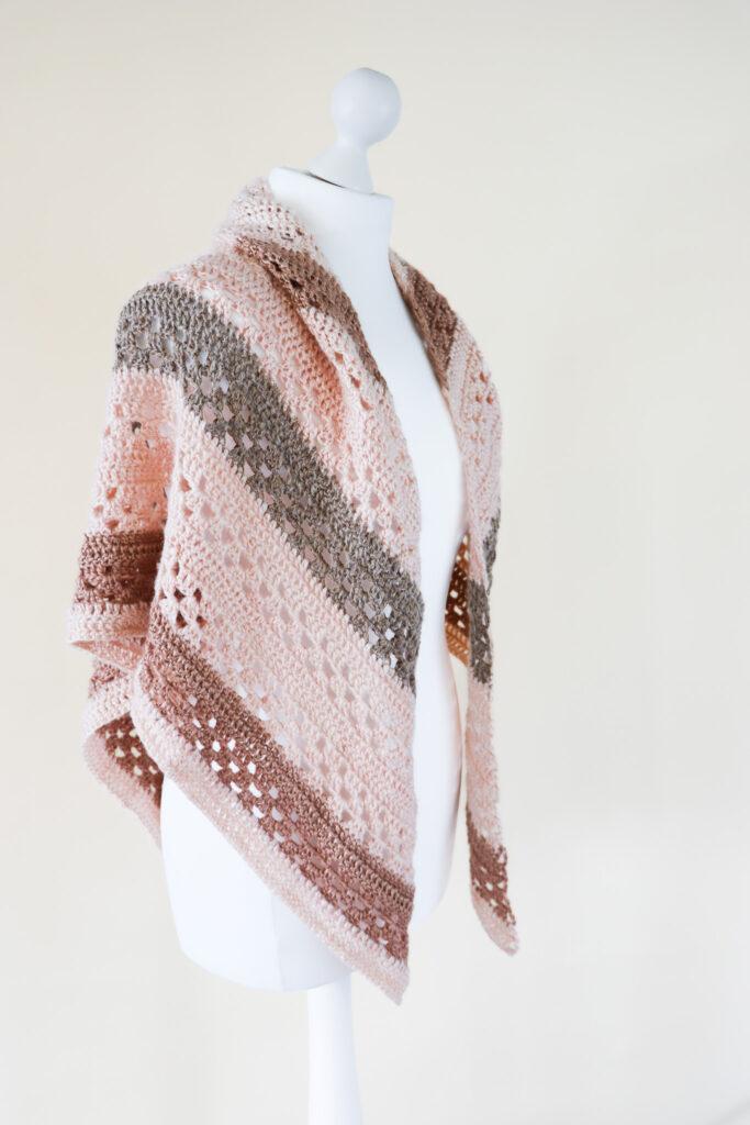 a crochet shawl made is neutrals shades of metallic yarn worn on a white mannequin stood in front of a cream background