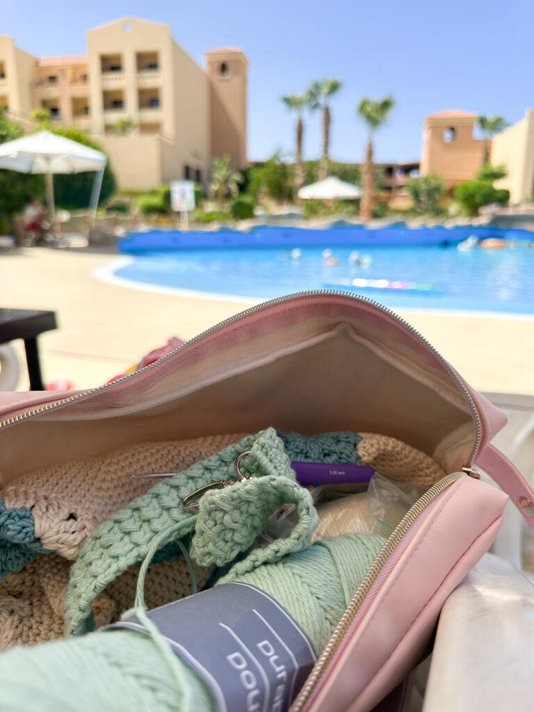 an image of a zip bag with a crochet hook, yarn and scissors inside with a swimming pool in the background. 