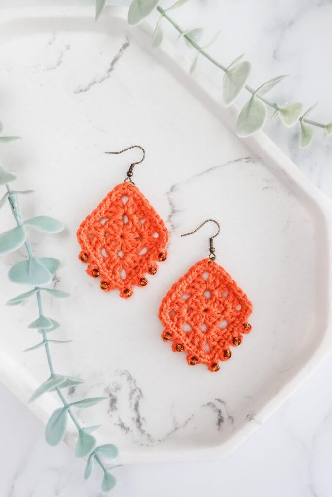 flat lay image of a pair of crochet earrings made in orange yarn lay on a white marble background with foliage in the top left corner.