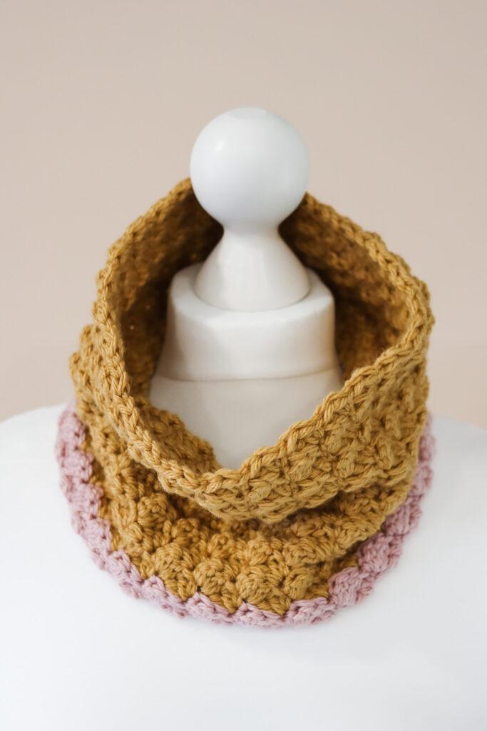 a textured crochet cowl in mustard and pink yarn modelled on a white mannequin torso in front of a pale pink backdrop.
