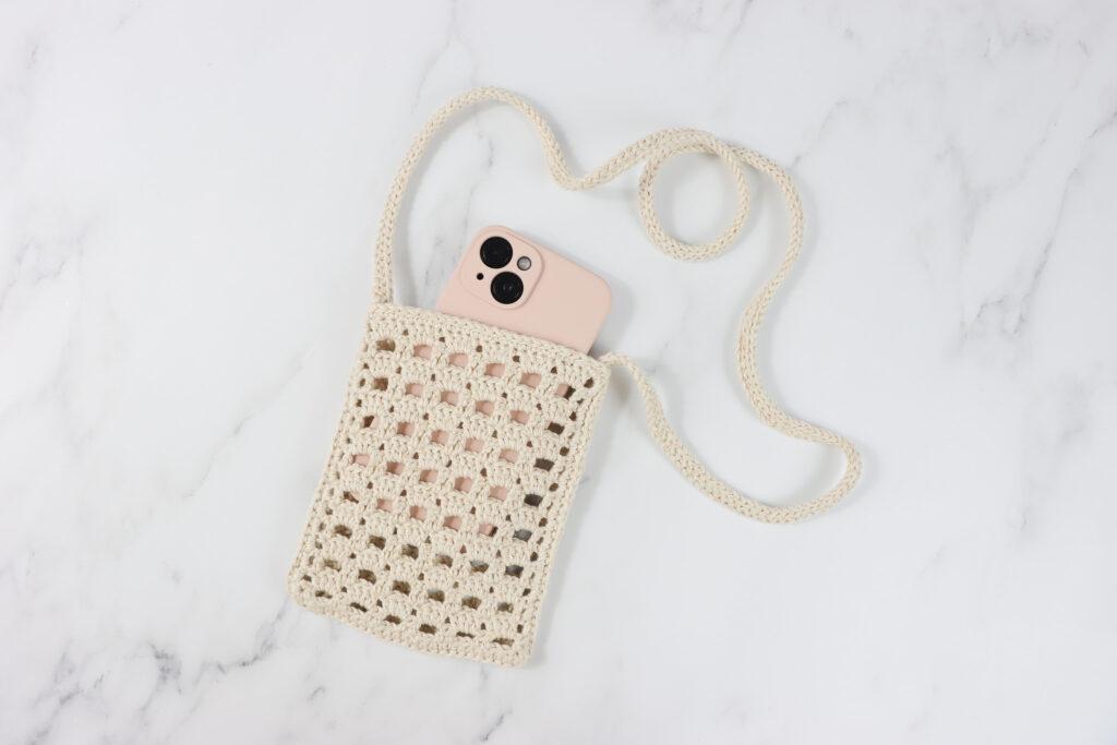 a crochet phone pouch made in a beige sparkly yarn lay flat on a marbled back ground with a pale pink iPhone inside.