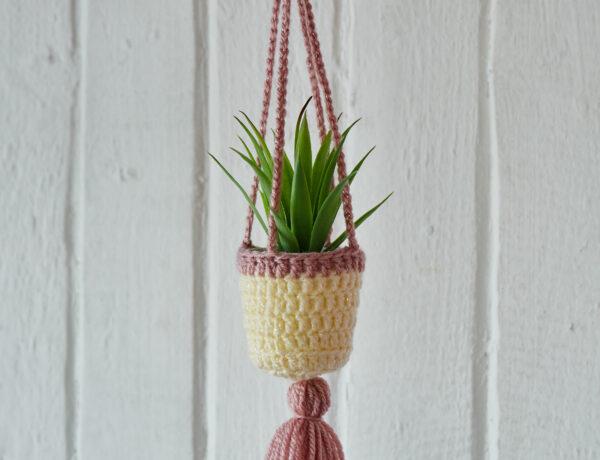 Crochet plant hanger made from cream and pink yarn with a plant inside, hanging against a white wood background.