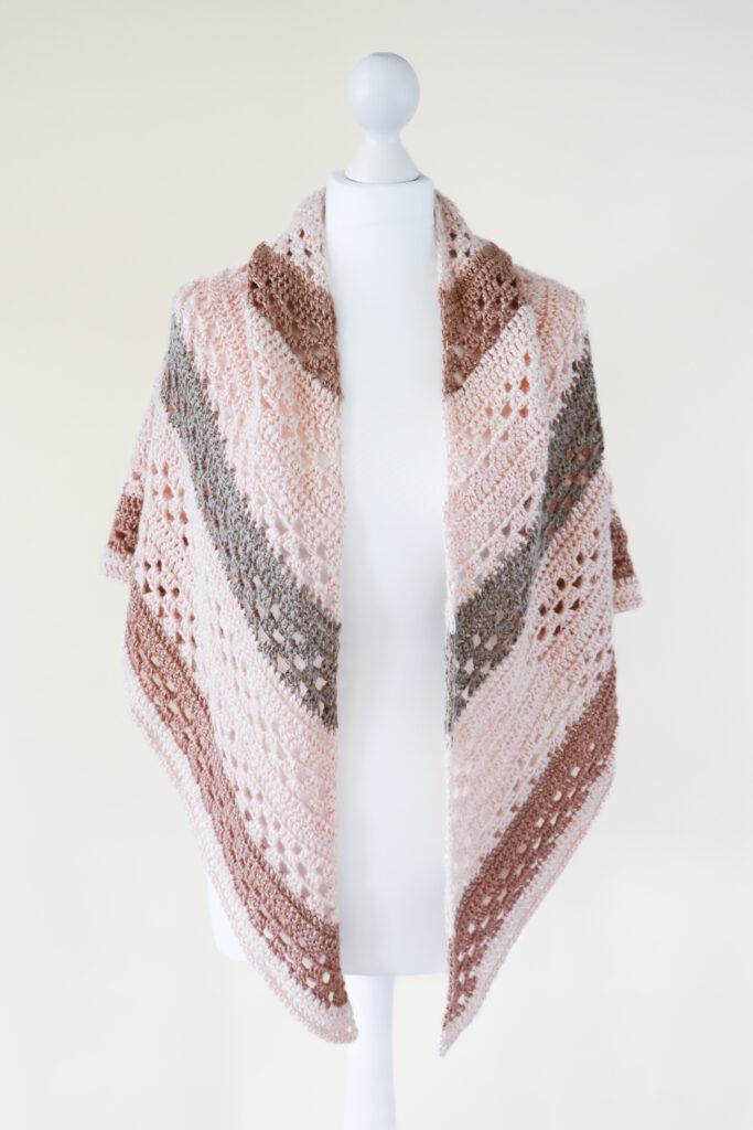 a crochet shawl made it pink and mauve metallic yarn displayed on a white mannequin intron of a cream background