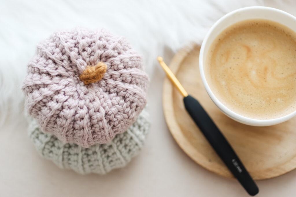 flay lay image of two crochet pumpkins stacked one alongside a cup of coffee and a black and gold crochet hook. 