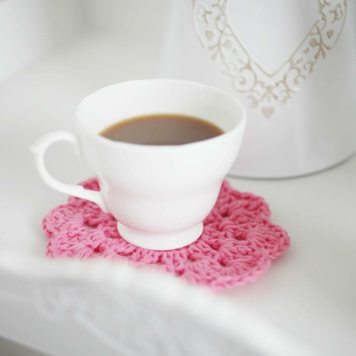 a close-up image of a white cup on a pink crochet coaster that is on a white tray.