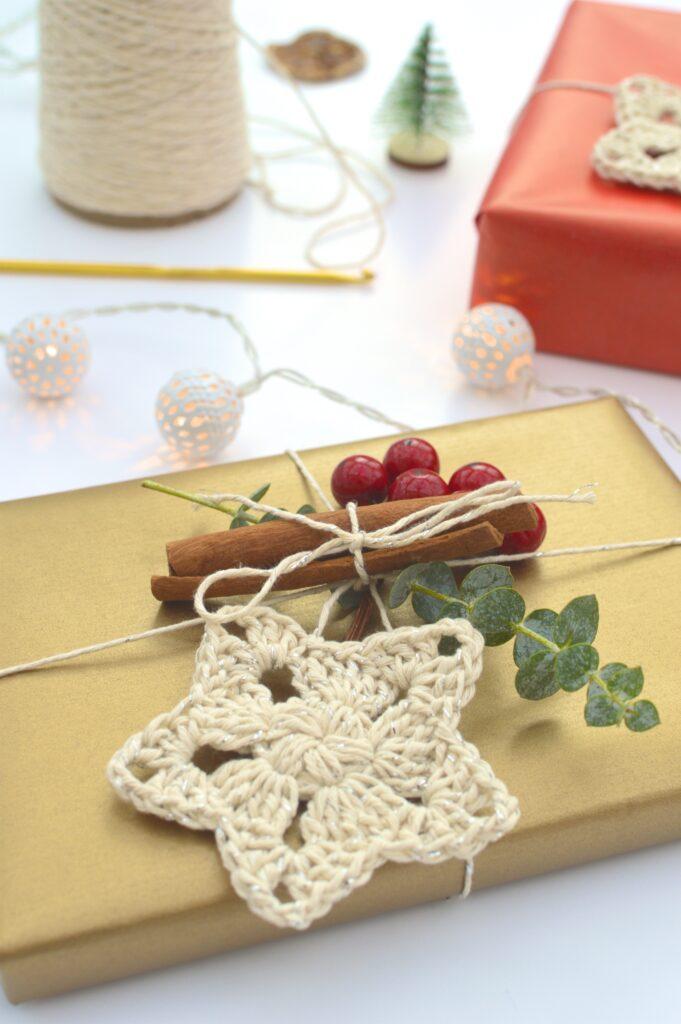 a crochet star attached to a present with twine, foliage and berries. In the background there is a crochet hook, twine and a mini Christmas tree