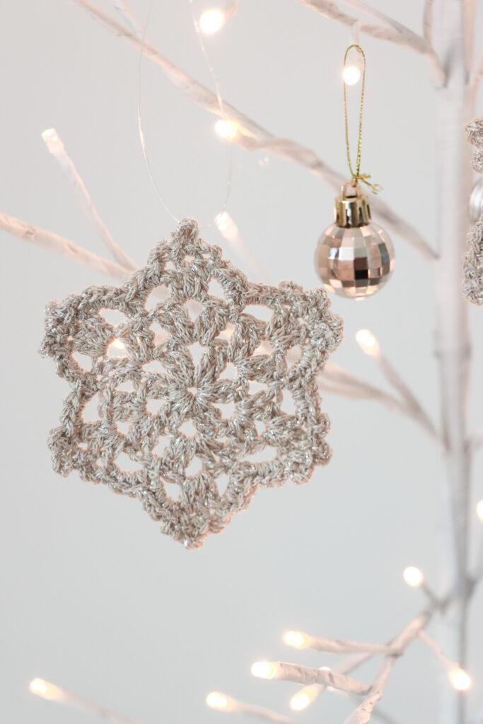 crochet snowflake made in gold sparkly yarn hanging from a white branch tree with lights on the end. 