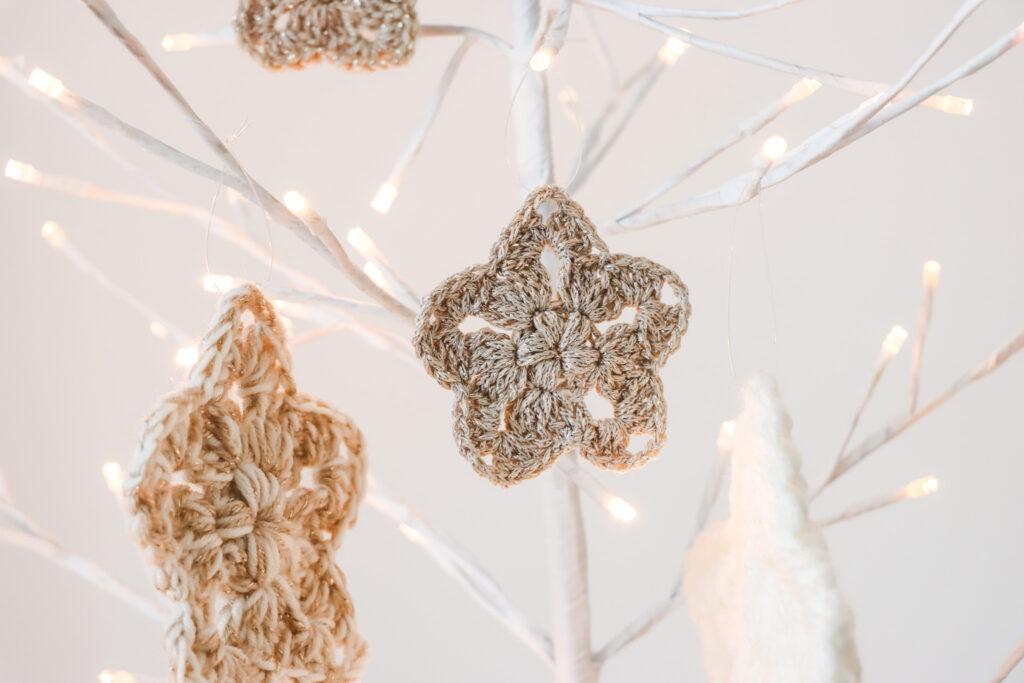 four crochet stars made in gold, cream and neutral sparkly yarn hanging from a white branch tree that has lights on the tips of the branches 