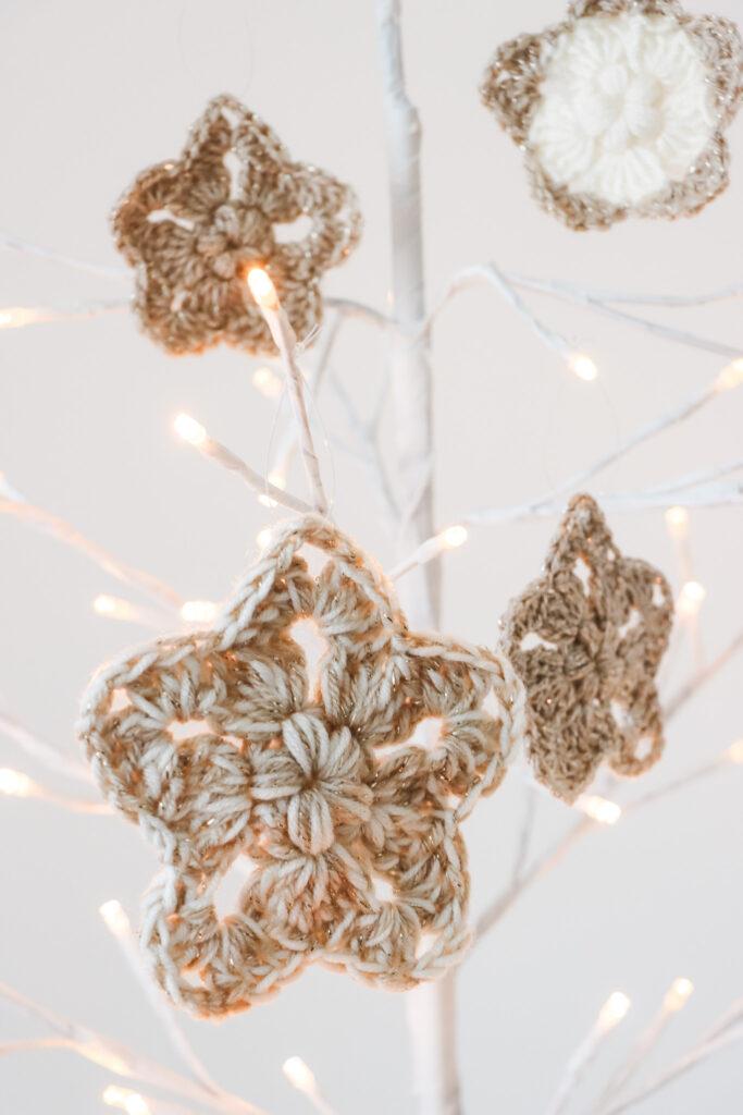 four crochet stars made in gold, cream and neutral sparkly yarn hanging from a white branch tree that has lights on the tips of the branches