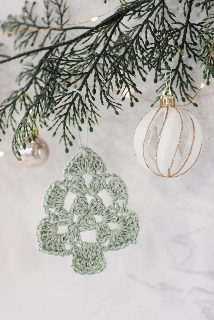 a crochet Christmas tree made in green yarn hanging from foliage with white and gold baubles hanging alongside