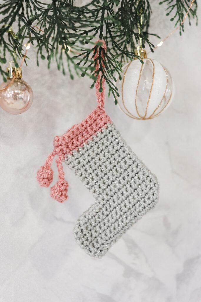 a mini crochet stocking made in sparkly grey and pink yarn hanging from foliage with white and gold baubles hanging alongside