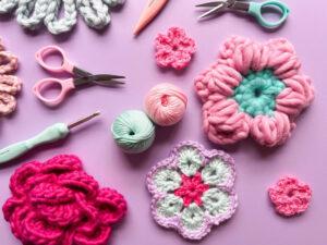crochet flowers, crochet hooks, yarn and scissors lay flat on a purple background.