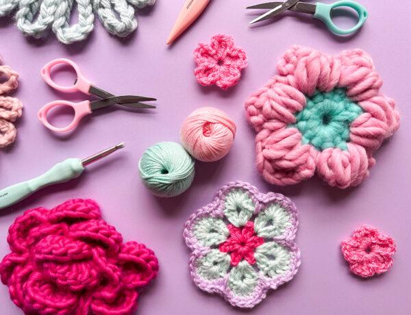 crocht flowers, crochet hooks, yarn and scissors lay flat on a purple background.