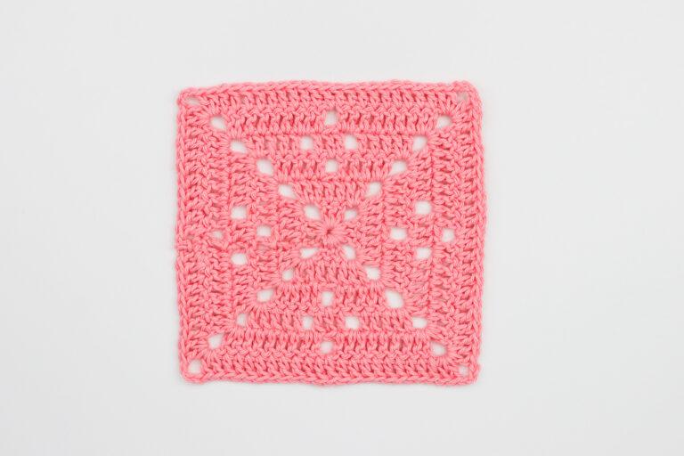 Pink filet granny square crochet motif displayed flat on a white background, featuring a lacy geometric pattern created with treble crochet and chain spaces.
