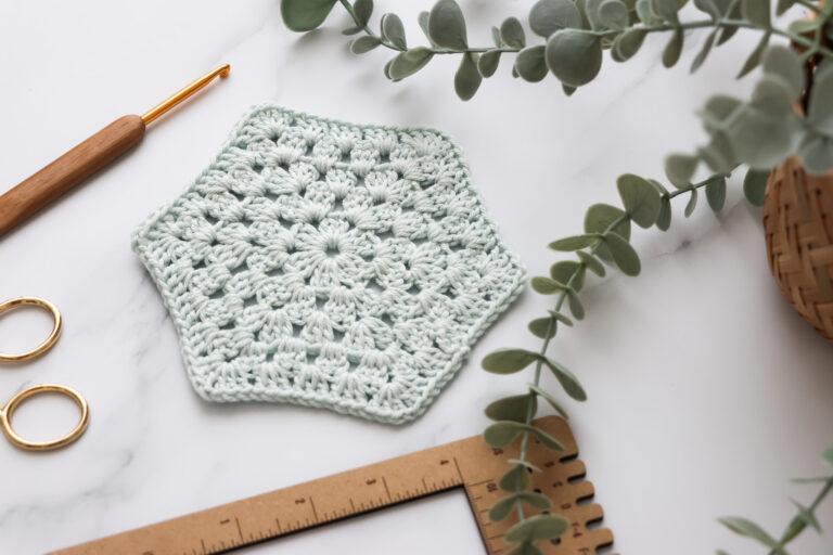 Flat lay of a light blue crochet hexagon motif made with puff stitches, surrounded by a wooden-handled crochet hook, gold stitch markers, a wooden ruler, and eucalyptus leaves on a white marble background.