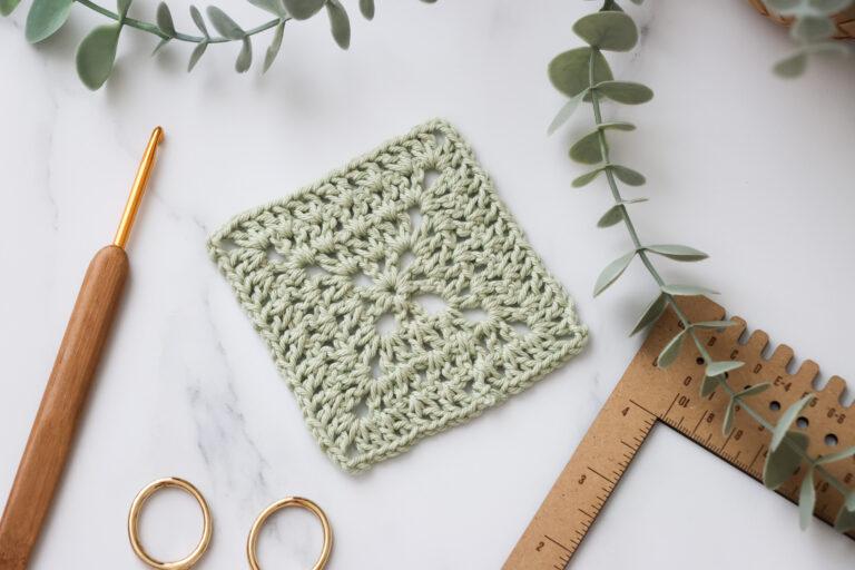 Light green crochet granny square made with soft cotton yarn, displayed on a white marble surface with a wooden-handled crochet hook, measuring tool, and eucalyptus leaves.