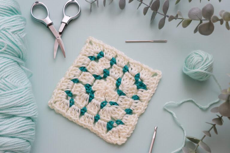 Crochet granny square in cream and teal yarn displayed on a light mint background, surrounded by crochet tools including scissors, a yarn needle, a crochet hook, and soft yarn balls.